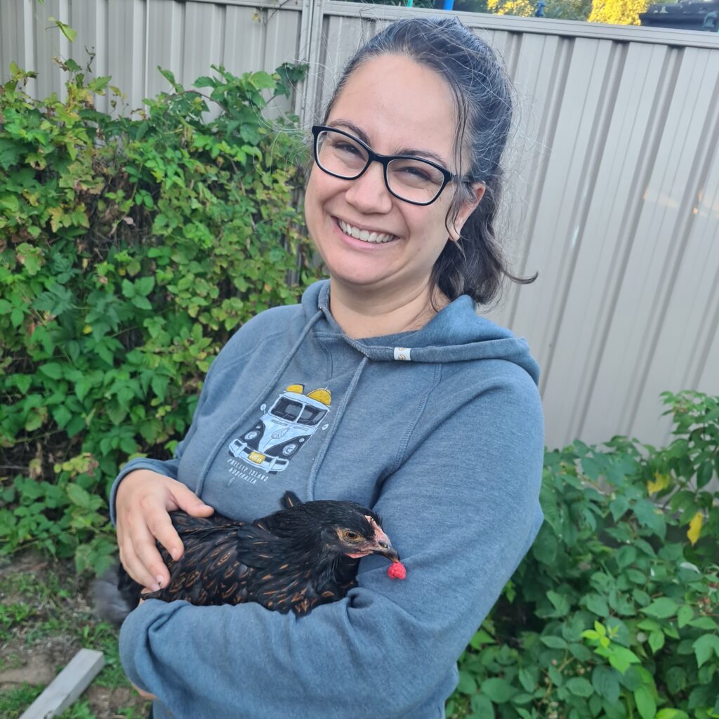 Amy in a blue hoodie holding a black chicken who is holding a raspberry in her beak. As you do, when you're a chicken.